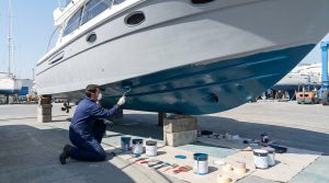 Choix et application de l'anti fouling pour bateau Homme masqué en combinaison appliquant de l'anti-fouling bleu sur la coque d'un bateau en cale sèche, outils au sol.