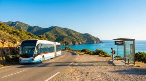 Bus en corse : réseau, tarifs et horaires officiels en 2026 Bus longeant la côte corse, mer bleue, montagnes. Un homme attend à un abribus moderne sous un ciel clair.
