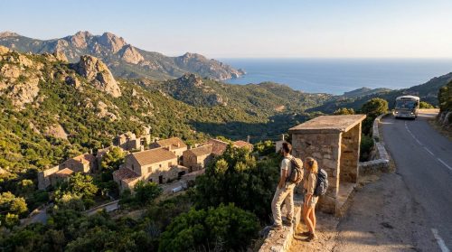 Deux randonneurs observent un village corse, la mer Méditerranée et des montagnes. Un bus roule sur une route sinueuse en Corse.