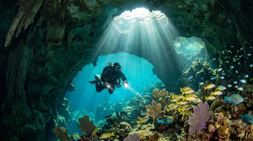 Un plongeur avec lampe explore une grotte sous-marine éclairée par un rayon de soleil traversant l'eau turquoise.